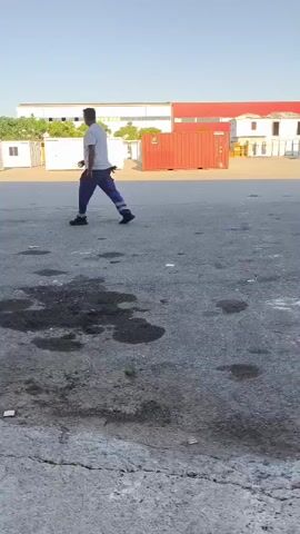 The video shows a person walking in an outdoor industrial area. The person is wearing a white t-shirt and blue pants. They are walking from left to right across the frame. In the background, there are several white modular buildings and a red shipping container. The ground is paved and appears to have some oil stains. The sky is clear and blue.