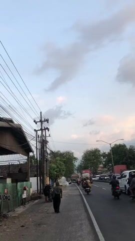A vertical, low-angle video shows a street scene in Kejapanan, Indonesia, at dusk. The sky is a pale blue with scattered, wispy clouds tinged with pink from the setting sun. Overhead, a dense network of power lines crisscrosses the frame, supported by utility poles.

On the left side of the street, a building with a corrugated metal roof and a light-colored wall is visible behind a green metal fence. Several people are walking along the sidewalk. In the foreground, a person in a patterned shirt and dark pants walks away from the camera. To their left, another person in a dark shirt and shorts stands near a parked motorcycle. Further down the sidewalk, two people are walking together, one wearing a red helmet.

The street itself is paved and has a white line marking the lane division. Traffic is moving on the right side of the road, primarily consisting of motorcycles. A few cars and trucks are also present, including a red shipping container on a truck in the distance. Lush green trees line the right side of the road, interspersed with various buildings and structures. The overall atmosphere is that of a typical evening street in a residential or semi-commercial area, with the gentle glow of twilight and the ambient sounds of daily life.