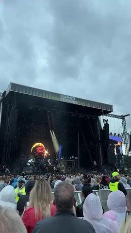 A wide shot shows a concert stage at the event "SUMMER SESSIONS". A large screen behind the stage displays a close-up of a man's face. Two spotlights are directed downwards from above. On the stage, several musicians are performing. In the foreground, a crowd of people is gathered, many wearing hooded jackets, suggesting cool or rainy weather. The sky is overcast.