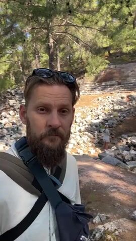 A man with a beard and sunglasses on his head films himself in front of ancient stone ruins. He is wearing a light-colored shirt and a backpack with a blue strap. The ruins appear to be an amphitheater, with tiered seating made of stone. Trees with green foliage are visible in the background, and there are scattered stones and debris around the site. The lighting suggests it is daytime.