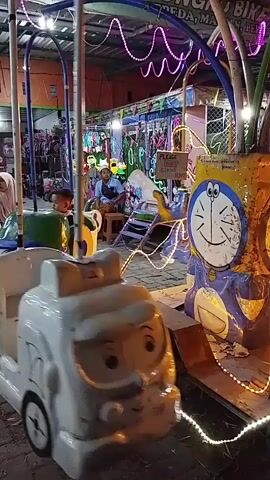 A child rides a carnival ride. The ride has colorful lights and characters, including Doraemon. There are other people in the background, and the ride appears to be in an outdoor market. The video is shaky, suggesting it was filmed on a mobile device.