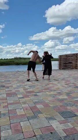 The video shows three people standing on a tiled patio near a body of water. The sky is blue with white clouds. In the foreground, a shirtless man in dark shorts is facing away from the camera, with his arms bent. To his right, a woman in a long black dress is facing him, with her arms raised. To the far right, another man in dark shorts is standing. The man and woman in the foreground appear to be interacting, possibly dancing or playing. The water is calm and there is greenery along the far bank. A wooden structure is visible on the right side of the frame.