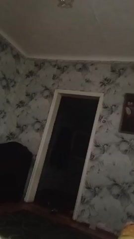 The media file captures an interior view of a room with floral wallpaper. The primary focal point is an open doorway, revealing a dark, unlit space beyond. A portion of a dark-colored piece of furniture, possibly a chair or sofa, is visible on the left side of the frame. On the right wall, a framed picture, likely religious in nature, is hung. The ceiling has a light fixture. The overall lighting suggests an indoor setting, possibly during the day or evening with interior lights on or off. The scene appears to be a residential room. There is no discernible activity or event taking place beyond the static view of the room. No text is visible in the media file.