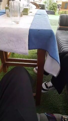 The video shows a table covered with a white and blue cloth. On the table, there is a glass, a tissue box, and some other objects. Under the table, there are people's legs. The person whose perspective the video is from is wearing black pants. Another person is wearing black pants and white and black sneakers. In the background, a person is walking away. The environment appears to be outdoors, with artificial grass on the ground and some furniture in the background.