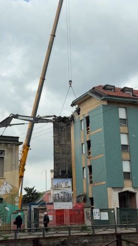 The video shows the demolition of a multi-story building. A large crane is lifting a section of the building's top floor, which has already been partially demolished. The building is light blue and tan, with a red tile roof. There are windows visible on the remaining floors. Below the building, a red fence runs along a grassy area. Two people are walking on the sidewalk in front of the fence. A sign displaying architectural renderings of a new building is positioned near the construction site. A water spray is visible on the left side of the frame, likely to control dust. The sky is overcast.