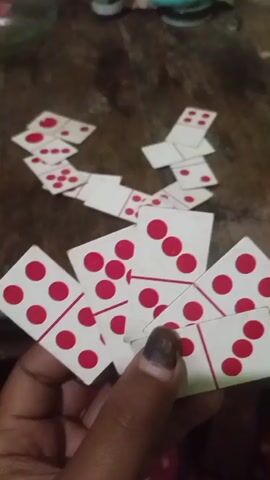The video shows a hand holding several dominoes. The dominoes are white with red dots. In the background, more dominoes are scattered on a wooden surface, arranged in a winding pattern. The lighting is dim, suggesting an indoor setting.