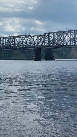 The video shows a wide, calm river with a large, metallic railway bridge spanning it. The bridge has a complex truss structure supported by two large concrete pillars in the water. The water's surface is rippled, reflecting the cloudy sky and the bridge. On the far bank, a lush green hillside is visible, covered with trees. The sky is filled with dramatic, overcast clouds, suggesting potentially stormy weather. A dark-colored train is seen moving across the bridge from left to right. There are no people visible in the video. The overall impression is one of industrial infrastructure set against a natural landscape under a moody sky.