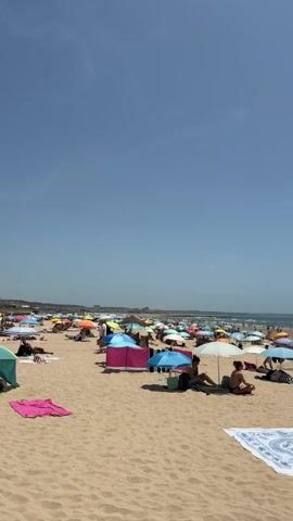 The video shows a crowded beach on a sunny day. Numerous colorful umbrellas are scattered across the sand, providing shade for beachgoers. People are seen relaxing on towels, sitting under umbrellas, and walking towards the ocean. The water appears calm with gentle waves, and several people are visible in the distance near the shoreline. The sky is clear and blue, suggesting pleasant weather conditions. The overall scene depicts a lively and popular beach destination.