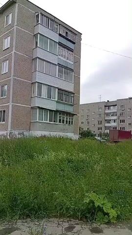This is a video clip showing a residential area in Nytva, Russia. The scene is dominated by large, multi-story apartment buildings, typical of Soviet-era architecture, with grey facades and numerous balconies. In the foreground, there is a patch of overgrown grass and weeds, with some concrete pathways.

The video pans across the area, revealing different aspects of the community. We see a playground with swings and other equipment, where a few children are present and actively playing. There's also a sports area with a basketball hoop and a soccer goal, suggesting a focus on outdoor recreation. A wooden bench is visible, offering a place to rest.

Further along, another apartment building is seen, and there are glimpses of trees and vegetation interspersed among the buildings, adding some natural elements to the urban landscape. In the background, a red truck is parked, and other vehicles are also visible, indicating a functioning residential environment.

The weather appears to be overcast and cloudy, contributing to a somewhat muted atmosphere. The time of day is difficult to pinpoint precisely but seems to be daytime, likely afternoon, given the lighting and the presence of children playing outdoors. There are no prominent emotional displays from the people visible; they seem engaged in their activities. No visible text is present in the video. The overall impression is a typical day in a Russian residential district, with community spaces being utilized.
