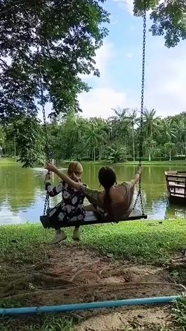 Two people are sitting on a swing hanging from a tree branch, swinging over a body of water. The person on the left has blonde hair and is wearing a dark floral dress and white sneakers. The person on the right has dark hair and is wearing a brown t-shirt and pants. The swing is moving back and forth. The background shows a calm lake surrounded by lush green trees and palm trees under a partly cloudy blue sky. A wooden platform or deck is visible on the right side of the lake. The foreground shows grass and dirt.