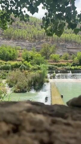 A scenic view of a body of water flowing over a series of small waterfalls, framed by lush green foliage in the foreground and a tree-covered hillside in the background. The water is a vibrant turquoise color, and the surrounding vegetation is dense and healthy. The scene suggests a natural, serene environment.