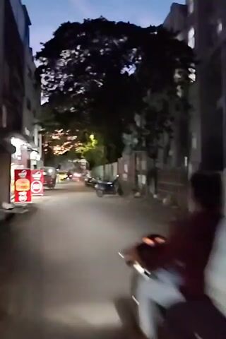 The video is a first-person perspective shot from a moving scooter or motorcycle on a narrow, paved street. The time of day appears to be dusk or early evening, with the sky showing a gradient of dark blue and some light. Streetlights and lights from buildings are visible. On the left side of the street, there is a building with a red and white sign that appears to be for a food establishment, possibly a burger joint, with an image of a burger. Further down the street on the left, there are more lights and some parked vehicles. On the right side, there are buildings with brick walls and some parked cars. The street is lined with buildings and trees. In the foreground, the right arm and shoulder of the rider are visible, wearing a maroon shirt. The rider is looking forward down the street. Other vehicles, including a car and possibly another scooter, are visible further down the street. The overall impression is of a residential or commercial street in an urban or semi-urban setting during twilight.