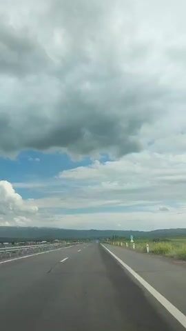 The video shows a view from the perspective of someone driving on a highway. The road is asphalt with clear white lane markings. The sky is filled with dramatic, gray clouds interspersed with patches of blue sky. In the distance, there are mountains and some signs of civilization, including buildings and a guardrail. The surrounding landscape appears to be rural, with green fields visible on the right side of the road. The sun is not directly visible, but the lighting suggests it is daytime, possibly overcast or a cloudy day. The overall impression is of a journey through a vast, open landscape under a dynamic sky.
