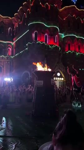 A nighttime outdoor event features a large bonfire in the center of the frame, with a crowd of people gathered around it. The bonfire is housed in a dark, cylindrical structure with some markings on it. Behind the bonfire, a building is illuminated with colorful lights, predominantly red and green, giving it a festive, castle-like appearance. The building has several windows and decorative architectural elements. Many people in the crowd are looking towards the bonfire and the building, some with their hands raised, suggesting they are actively participating in the event. Several individuals in the crowd appear to be wearing reflective vests, possibly indicating they are staff or organizers. To the right of the bonfire, a person dressed in a red and black costume stands on a small stool, facing the crowd and appearing to be performing or leading a segment of the event. There is a general sense of celebration and engagement among the attendees. The ground in front of the bonfire appears wet and reflective, possibly from recent rain or water features.