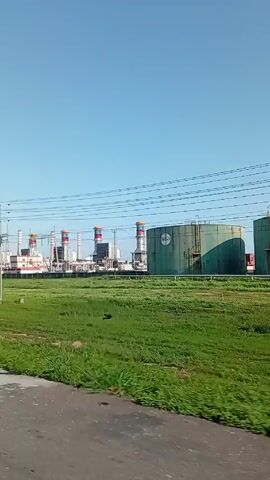 The video shows a wide shot of an industrial facility on a sunny day. The facility consists of various structures, including tall stacks with red, yellow, and blue tops that appear to have the colors of the Venezuelan flag. Other structures suggest a power plant or refinery, with metal frameworks and tanks. There are large, cylindrical tanks painted a light green color. These tanks have labels on them. The facility is situated in a relatively flat, open area with a mix of green grass and patches of bare earth. Overhead, there are numerous power lines strung between utility poles. The camera appears to be in a moving vehicle on a paved road. The sky is clear and blue. The video does not contain any human figures or interactions.