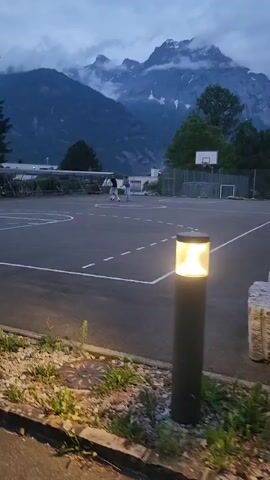 The video shows a basketball court with two people playing in front of a mountainous backdrop. The court is dark asphalt with white lines demarcating the boundaries and key areas. A basketball hoop stands at one end, and there is a fenced-off area behind it. Bicycles are parked beneath a shelter to the left of the court. The people, who are small and distant in the frame, are moving around the court, possibly playing basketball. The environment is marked by a large mountain range with visible snow and cloud cover. There are trees and greenery scattered around the area. A lamppost stands in the foreground, its light casting a warm glow on the rocks and vegetation at its base. The sky is overcast. The camera slowly pans to the left, keeping the basketball court and mountains in view.