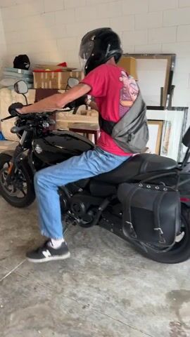 A person wearing a red t-shirt, blue jeans, a black motorcycle helmet with a clear visor, black gloves, and a gray crossbody bag is sitting on a black motorcycle. The person is in what appears to be a garage or workshop, with a concrete floor and a white brick wall in the background. There are various objects visible in the background, including boxes, furniture, and what looks like a framed picture. The person is looking towards the camera and their right hand is on the handlebars of the motorcycle. Their left hand appears to be resting on the seat of the motorcycle. The motorcycle has saddlebags on either side.