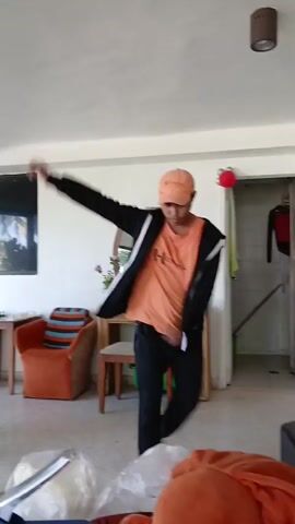 A young man wearing an orange t-shirt, a black zip-up hoodie, black pants, and an orange baseball cap is dancing in a room. He is performing various arm movements and spins. The room appears to be a living area with orange chairs, a wooden dresser with a mirror, and a doorway in the background. There are also some plastic bags and a speaker visible in the foreground.