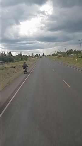 The video captures a journey along a two-lane asphalt highway, presumably in a rural area of Bomet, Kenya, from the perspective of a moving vehicle. The sky is heavily overcast with dark grey clouds, indicating a cloudy day, possibly before or during a rain shower, as a distant section of the road appears hazy and wet.

Various vehicles are seen on the road. Initially, a motorcyclist is visible on the left shoulder. As the recording vehicle proceeds, other traffic comes into view. A white passenger van is parked on the right side of the road with several people gathered around it. A large white truck, various cars (including sedans and dark grey SUVs), and another motorcyclist pass by. A prominent feature is a red tractor slowly moving on the left, pulling a large blue cylindrical water tank. Utility poles line both sides of the road, and the surrounding landscape consists of grassy areas, scattered trees, and distant, simple buildings. The activity primarily involves vehicles in transit on the highway. No distinct textual information is clearly readable.