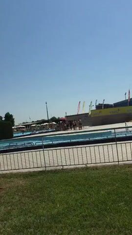 This video shows a sunny day at a water park, likely in Reggio nell'Emilia, Italy. The scene is dominated by a swimming pool with several people enjoying themselves. Many are floating on yellow inflatable rings in the blue water. In the background, there's a large stadium structure, suggesting the water park might be adjacent to a sports facility. A red and white umbrella with the word "AQUATICO" printed on it is prominently displayed in the foreground, providing shade. Flags with various designs are visible further in the background, adding to the festive atmosphere of a public leisure space. The clear blue sky and bright sunlight indicate fair weather, likely midday or early afternoon. The overall mood appears relaxed and recreational, with families and individuals engaging in typical water park activities.