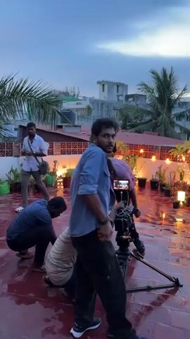 The video captures a film or video shoot on a red-tiled rooftop at dusk or early evening. Several crew members are visible, engrossed in their tasks. A camera is set up on a tripod, with one person operating it. A man in a light blue shirt stands prominently, initially looking towards the camera, then focusing on a smartphone, likely monitoring the shoot. In the background, a woman in a sari stands, facing away from the camera, positioned for a shot amidst potted plants and decorative string lights that are illuminated. The rooftop floor appears wet, possibly from rain or having been intentionally watered. The sky is overcast, transitioning from daylight to night, and other urban buildings and palm trees are visible in the background.
