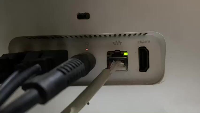 The video shows a close-up shot of the rear ports of a white electronic device, likely a router or a gaming console. Several cables are plugged into the ports. On the left, there are at least two black cables connected to ports that are not fully visible. In the center, an Ethernet cable is plugged into an Ethernet port, with its connector housing visible. The Ethernet port has two indicator lights, a green light which is illuminated, and an amber light which briefly flashes on and then off. To the right of the Ethernet port, an HDMI port is visible but empty. Above the Ethernet port, a small red indicator light is illuminated on the device itself. The device has a textured surface with ventilation slits along the top edge. The background appears to be a plain, light-colored surface. The lighting is dim, suggesting an indoor environment. There are no people visible in the video. The actions depicted are static, with the focus on the connected ports and indicator lights.
