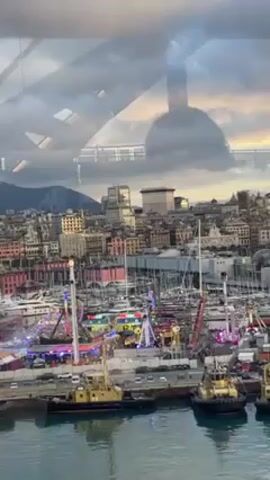 The video shows a cityscape with a carnival or fair in the foreground. There are various rides and attractions visible, lit up with colorful lights. In the harbor, several boats and tugboats are docked. The buildings in the background vary in height and architecture. A mountain range is visible in the distance. The sky is overcast with a hint of sunset. The view appears to be taken from inside a building or structure, as portions of the structure’s framework are visible, reflecting off the glass. An indiscernible child’s voice can be heard.