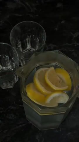 The video shows a close-up shot of a glass pitcher filled with water and lemon slices. A hand is visible, using a spoon to press the lemon slices against the side of the pitcher, presumably to extract their juice. Two empty glasses are positioned to the left of the pitcher. The background appears to be a dark, textured countertop.