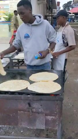 Here is a forensic description of the video:

The video shows two men cooking flatbreads on an outdoor griddle. The griddle is heated by a fire contained within a rusty metal enclosure. The man in the foreground is wearing a gray hoodie and jeans, and is using a spatula to flip the flatbreads as they cook. He also appears to be adding oil from a small bottle. A second man, wearing a white apron and a baseball cap, stands behind him, potentially assisting.

Behind the griddle, there are two tables: one with a stack of cooked flatbreads, and another with unbaked dough balls. A man sits at the second table, appearing to be preparing more dough. A pharmacy with a glass facade is visible in the background. A customer in a pink dress approaches the flatbread stand to make a purchase.