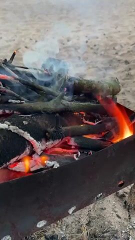 A close-up shot of a small, portable barbecue grill with burning wood and branches on a sandy beach. Smoke rises from the fire, and flames are visible. In the background, the sandy beach stretches towards the ocean, and a person is faintly visible in the distance. The overall scene depicts an outdoor fire being prepared on a beach.