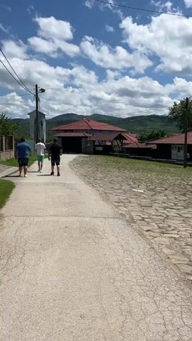 The video opens on a bright, sunny day in Rayuvtsi, Bulgaria, showing three men walking away from the camera along a concrete road. To their right is a cobblestone path. The men are casually dressed in shorts and t-shirts. In the background, traditional houses with red tile roofs and dark wooden features are nestled among green trees, with verdant, forested hills rising in the distance under a blue sky adorned with white, fluffy clouds.

The camera then pans right, revealing more of the scenic village. A stone wall topped with a black metal fence delineates a well-maintained grassy area where a flagpole stands with an indistinct flag. Several white, tent-like structures or gazebos are visible within this area, suggesting an outdoor event or dining space. Beyond these, more traditional Bulgarian houses with white walls and red roofs are seen, surrounded by greenery. The panoramic view emphasizes the picturesque rural setting, characterized by its traditional architecture and natural landscape. No discernible text is visible on any signs or objects throughout the video.