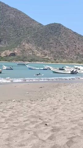 Here is a description of the video:

The scene is a beach with a view of a large mountain in the background on a sunny day.  The beach has light brown sand with gentle waves lapping at the shore.  Several small boats are floating in the water near the shore. A person is lying on a surfboard in the water, paddling with their hands.  The water is a light turquoise color.