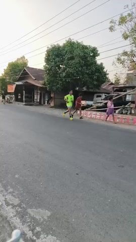 The video shows a street scene. There are several people running and riding motorcycles. A white SUV is also visible. The buildings lining the street appear to be shops or houses. There are trees and utility poles along the street. The sky is overcast, suggesting either early morning or late afternoon. The road surface is asphalt with some visible wear and tear. The overall atmosphere appears to be casual and active.