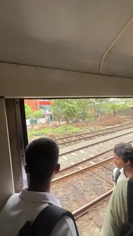 Two young men are looking out the window of a train carriage. The man on the left is facing away from the camera, his back to the viewer, wearing a white polo shirt and a black backpack. The man on the right is partially visible from the chest up, looking out the window. He is wearing a white shirt and has dark curly hair. Outside the window, train tracks are visible, with green foliage and some buildings in the background. The train appears to be moving, as indicated by the blurred motion of the scenery outside. The interior of the train carriage is visible at the top of the frame, showing the ceiling and the window frame.