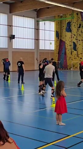 Here is a plain text description of the video:
The video shows a group of people training in what appears to be a martial art or combat sport, inside a gym. The gym has a blue floor marked with yellow lines, and there are yellow cones scattered around the training area. Several men, some wearing protective gear such as headguards and shin guards, are practicing techniques with boxing gloves, under the supervision of instructors. One instructor is wearing a grey shirt and appears to be demonstrating moves to a trainee. In the foreground, a woman with red hair and a red dress is seen observing the training. In the background, there is a climbing wall. Other people are also seen watching the training session.