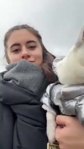 A young woman is holding a small, white dog wearing a silver puffer jacket. She is wearing a black puffer jacket and a grey scarf. She is smiling and looking at the camera. The dog looks to the side. Behind them is a cloudy sky, trees, and a Ferris wheel.
