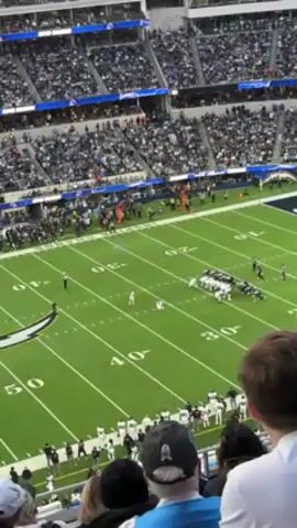 The video shows a football game in progress from an elevated position in the stands. A kicker in a white uniform attempts a field goal. A large crowd is seated in the stadium. The field is green with white yard markers and the team logo painted on it. The goalposts are visible in the background, and the kick is successful. The video is slightly shaky.