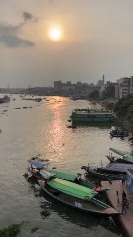 The video pans across a busy river scene during sunset. A golden sun hangs low in a hazy sky, casting a shimmering reflection on the water. Several boats of various sizes are visible. In the foreground, two green-canopied boats are docked, with a few people standing or sitting in them. Another small boat with a blue canopy is tied up next to them, also with people on board. Further out in the river, more boats, including a larger green ferry-like vessel, navigate the water. A distant city skyline with buildings and a minaret is visible on the horizon. People are seen on a dock to the right of the frame, some walking and others standing near the boats. The water is murky and appears to have some aquatic vegetation floating on its surface.