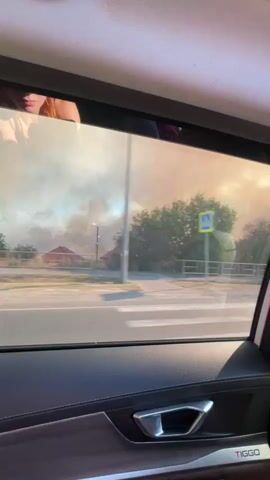 The video is filmed from inside a vehicle, looking out the passenger window. The window is tinted. Outside, a road can be seen with trees and some buildings in the background. A white car is driving on the road. There is a large plume of smoke in the distance, creating an orange haze in the sky. The interior of the car is visible, including the door panel with a handle and the "TIGGO" logo. A person's head is reflected in the top of the window. The sky is bright, suggesting daylight.