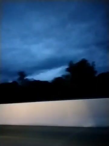 The video shows a dark, cloudy sky, likely at dusk or during a storm. Silhouetted trees are visible along the horizon, with a blurry, white surface in the foreground that could be part of a car's dashboard or a road barrier. The camera appears to be moving, suggesting the footage was captured from a vehicle. There are no people or discernible actions visible.
