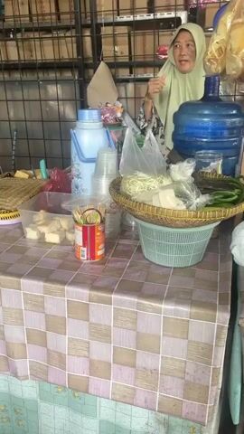 A vendor, a woman wearing a headscarf, stands behind a table laden with food items. To her left, on the table, are containers of what appear to be food ingredients, possibly for a meal. Among these are cubed items, plastic bags filled with chopped vegetables, and skewers. To her right, a large blue water dispenser is visible. In front of the dispenser, a woven basket holds more fresh ingredients, including green peppers and what looks like tofu. The table itself is covered with a checkered tablecloth. Behind the vendor, shelves are stocked with various packaged goods. To the right of the table, more shelves hold an array of products, some of which are brightly colored. The ground beneath the table is concrete. Further to the right, a collection of plastic stools are scattered around. The overall impression is of a small food stall or market stall, possibly outdoors given the ambient light.