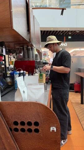A young man wearing a beige bucket hat, black t-shirt, and dark jeans stands behind a coffee bar in what appears to be a shopping mall. He is operating a coffee grinder, pouring coffee beans into it from a yellow bag labeled "MOKORO". The coffee bar is made of wood with visible storage compartments and features a variety of coffee cups, milk cartons, and a red espresso machine. A white bag with a palm tree logo is also visible on the counter. The mall environment is bright, with a few distant figures and retail signage visible in the background. The floor has a brown and orange tiled pattern.