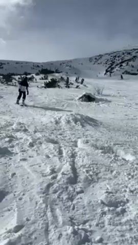 The video shows a person snowboarding down a snow-covered mountain slope. The person is wearing a black and white jacket, black pants, and black boots. They start from a higher point on the slope and snowboard in a curving path downwards. The landscape includes snow-covered terrain, scattered small trees, and other skiers visible in the distance. The sky is partly cloudy. The camera follows the snowboarder's descent.