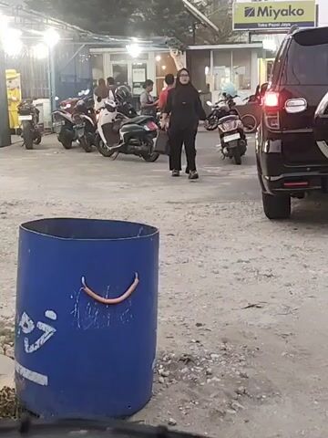 At 00:00, a woman wearing a black hijab and a black abaya walks towards a black SUV parked in a lot. She carries a black handbag in her left hand. In the background, several motorcycles are parked, and a building with lights on is visible. A blue trash can with a smiley face drawn on it is in the foreground. At 00:01, the woman reaches the SUV and opens the rear passenger door. At 00:02, she places her handbag on the seat inside the SUV and then closes the door. At 00:03, the woman walks away from the SUV, still in the same clothing. At 00:04, she is seen walking further away from the vehicle. The scene appears to be an outdoor parking area, possibly near a shop or restaurant.