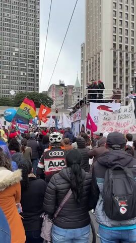A crowd of people marches down a city street flanked by tall buildings. Some people are carrying flags and signs, most in Portuguese. A double-decker trailer moves slowly through the crowd. People standing on the top deck are holding signs and banners. One man holds a piece of paper and addresses the crowd with a microphone. The weather is overcast.