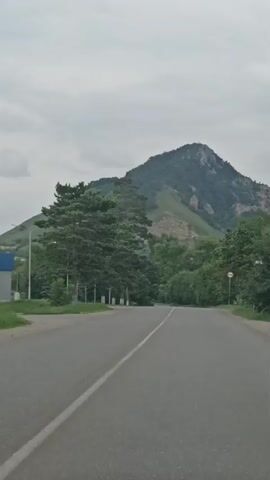 The video begins with a view of a paved road leading towards a mountain. On the left side of the road, there are trees and a fence. On the right side, there are more trees and a circular red and white sign. The sky is overcast. The camera appears to be moving forward along the road. The mountain is covered in greenery and has rocky outcrops. The overall scene is rural and natural.