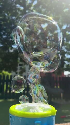 A bubble machine is in the foreground, emitting a stream of large, iridescent bubbles into a sunny outdoor environment. The background features a wooden fence, green grass, and a large tree with lush foliage. A swing chair is visible hanging from the tree branch. The camera focuses on the bubbles as they rise and float away. The lighting suggests it is daytime, possibly late morning or early afternoon.