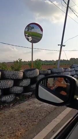 The video begins with a shot of a wall made of stacked tires, some painted white, bordering a rural road. In the side mirror of a vehicle, a person wearing a hijab is visible holding a phone, possibly taking a selfie. The vehicle is moving, indicated by the blurring of the scenery. Beyond the tire wall, there are fields with crops and trees under a clear sky with scattered clouds. A utility pole is also visible. The camera pans to the right, revealing more of the rural landscape and the continuation of the tire wall. The vehicle appears to be traveling on a paved road.