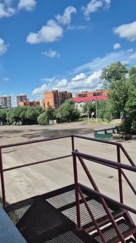 A black car drives across a paved area in front of buildings with red roofs and trees. The sky is blue with white clouds. A metal staircase with red railings is in the foreground.