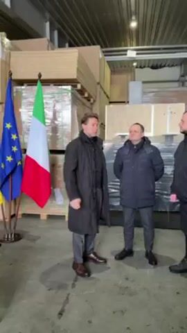 In an industrial-looking space with a light gray floor and a high corrugated metal ceiling, three men stand near flags and a desk.

The flags of the European Union, Italy, and Ukraine are arranged on stands behind the desk. Behind the men, stacks of wooden pallets and wrapped packages are visible.

The man on the left wears a long black coat, gray trousers, and brown shoes. He is gesturing with his hands as he speaks.

The man in the center wears a dark blue puffer jacket, dark gray trousers, and black shoes. He stands with his hands behind his back, listening.

Only the rightmost man's arm is visible. He appears to be wearing a jacket similar to the man in the center.

The light in the space is bright and uniform.